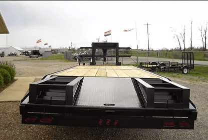 Black flatbed trailer parked on wet pavement with flags and industrial yard in the background