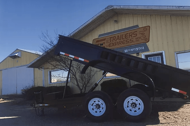 Black dump trailer with raised bed parked outside a tan trailer building.