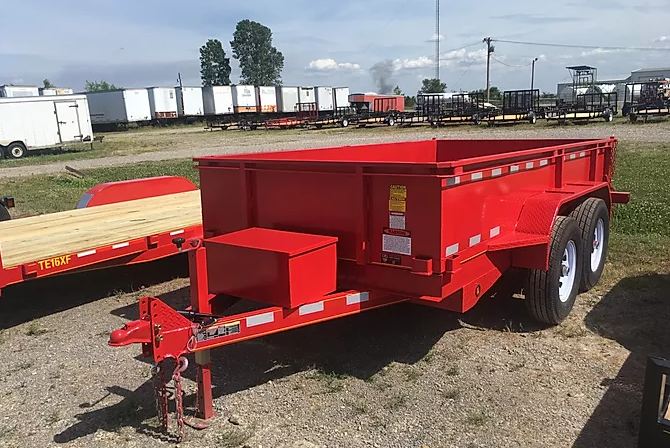 Red flatbed utility trailer with tandem wheels and hitch parked on a gravel lot near rail cars