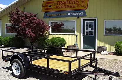 Small black utility trailer parked in front of a trailer shop sign with trees and a yellow building behind it