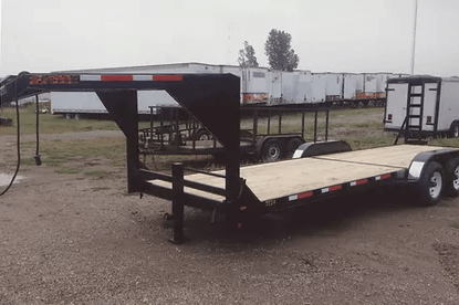 Black flatbed trailer with raised front platform parked on gravel lot, with white trailers in the background