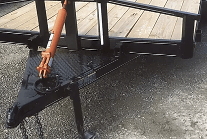 Black trailer hitch coupler with orange jack on a wooden deck beside pavement