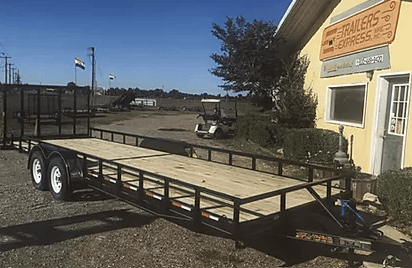 Black utility trailer parked beside a building on a gravel lot under clear blue sky