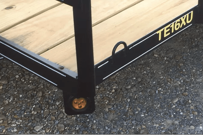 Black metal trailer frame labeled “TE16XU” on gravel beside a wooden deck