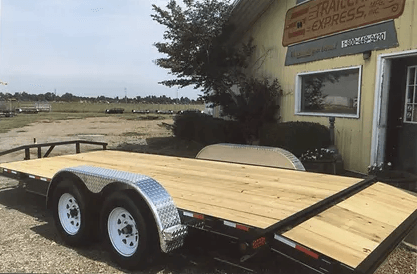 Wooden flatbed trailer with dual axles parked beside a building under cloudy skies.