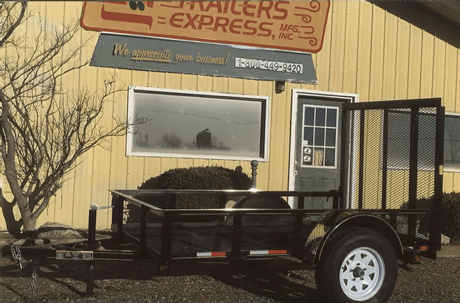Black utility trailer parked in front of a yellow building with a sign and window.