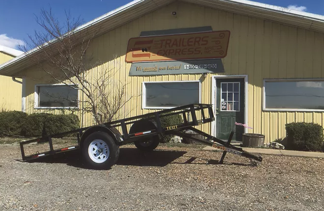 Small utility trailer parked outside a yellow building with signage and a door, on a gravel lot.