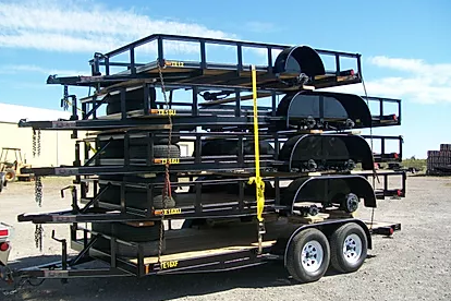 Stacked black metal trailer frames on a dual-axle utility trailer in a gravel lot