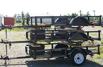 Stacked black utility trailers in a gravel yard under a blue sky.