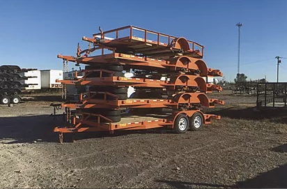 Stacked orange metal frames on a trailer in an open gravel lot under a blue sky.