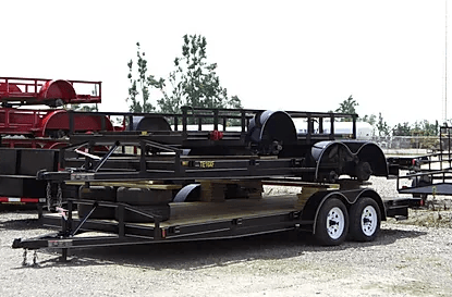 Black flatbed utility trailer with dual white wheels parked on gravel, with stacked equipment behind it