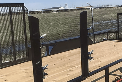 Outdoor wooden deck with black metal railing beside a grassy field and blue sky.