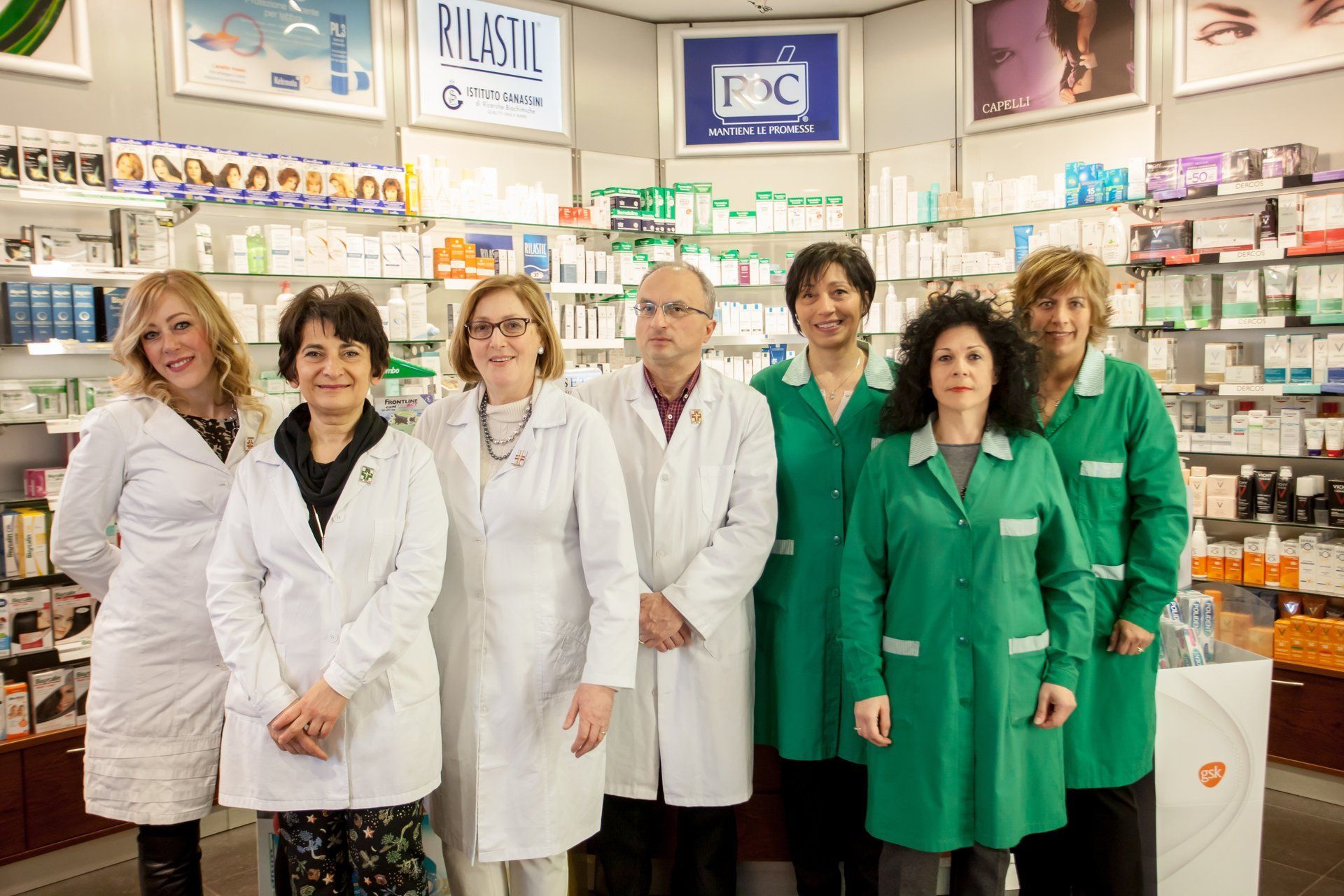 Un gruppo di sette membri dello staff della farmacia, in camice bianco o verde, sono in fila dietro il bancone.