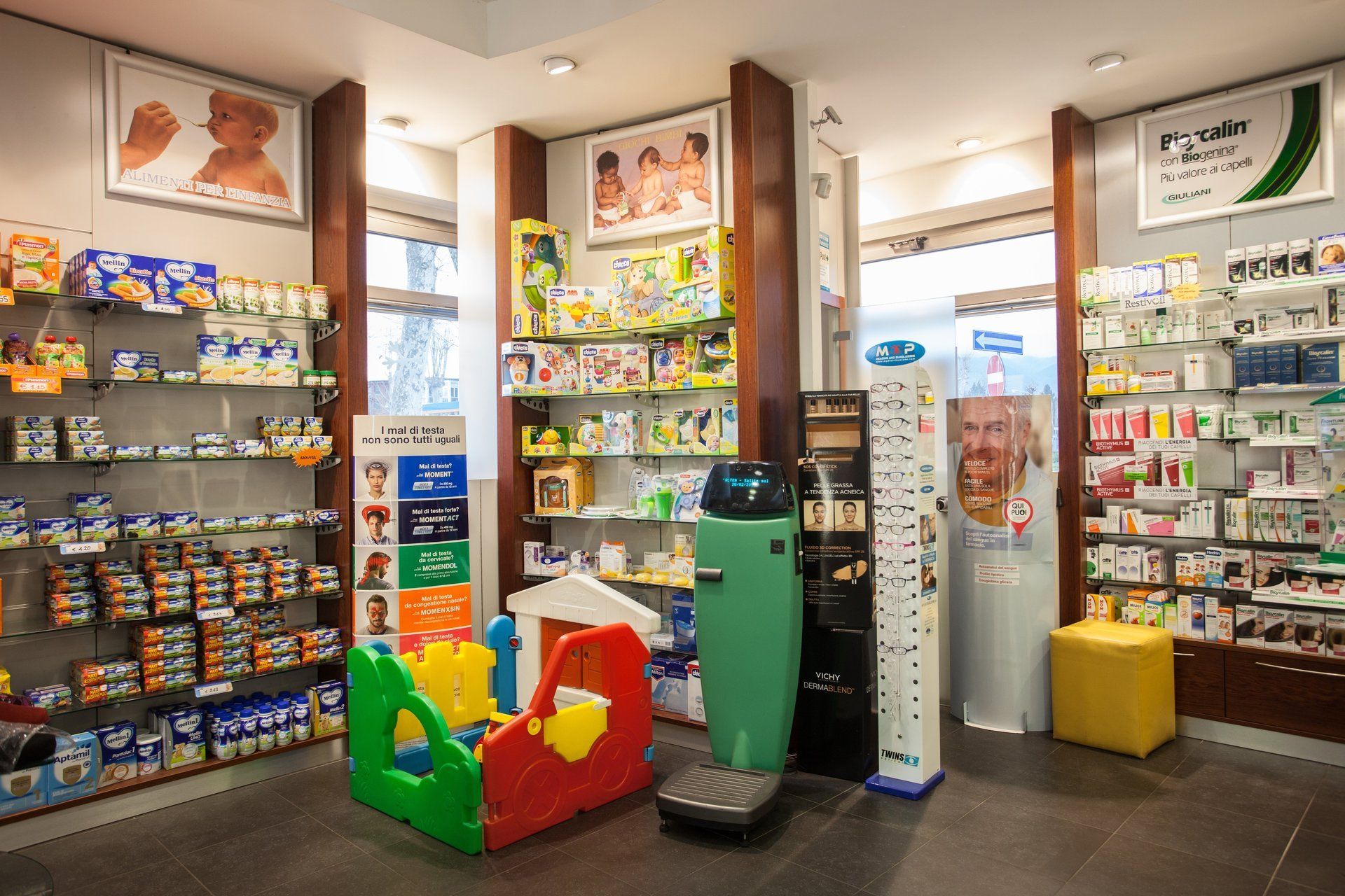 L'interno di una farmacia con scaffali pieni di prodotti per neonati, un'area giochi in plastica colorata, una bilancia e poster sulla salute.