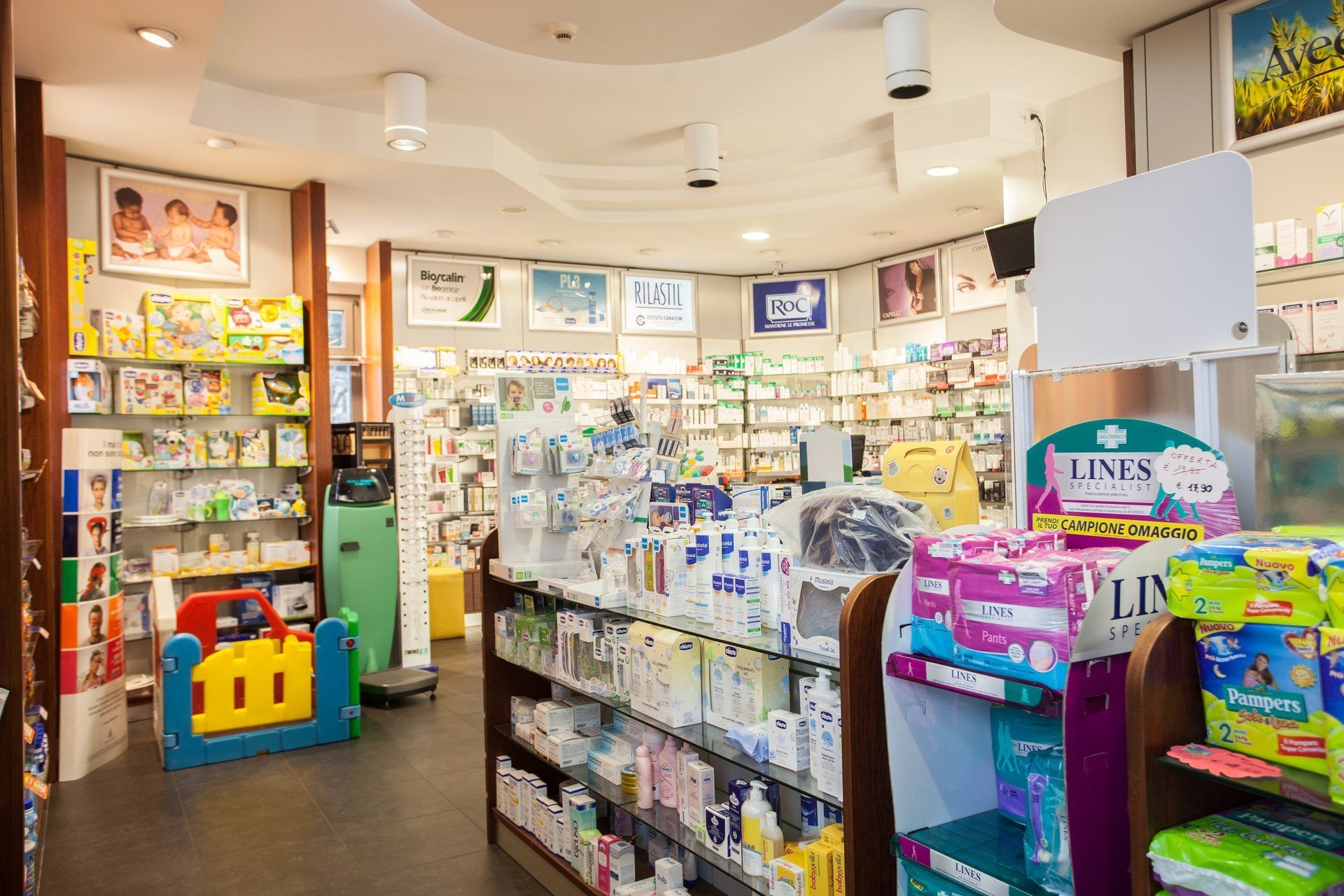 L'interno di una farmacia ben illuminata, con scaffali pieni di prodotti per la cura della persona, articoli per neonati e una colorata area giochi.