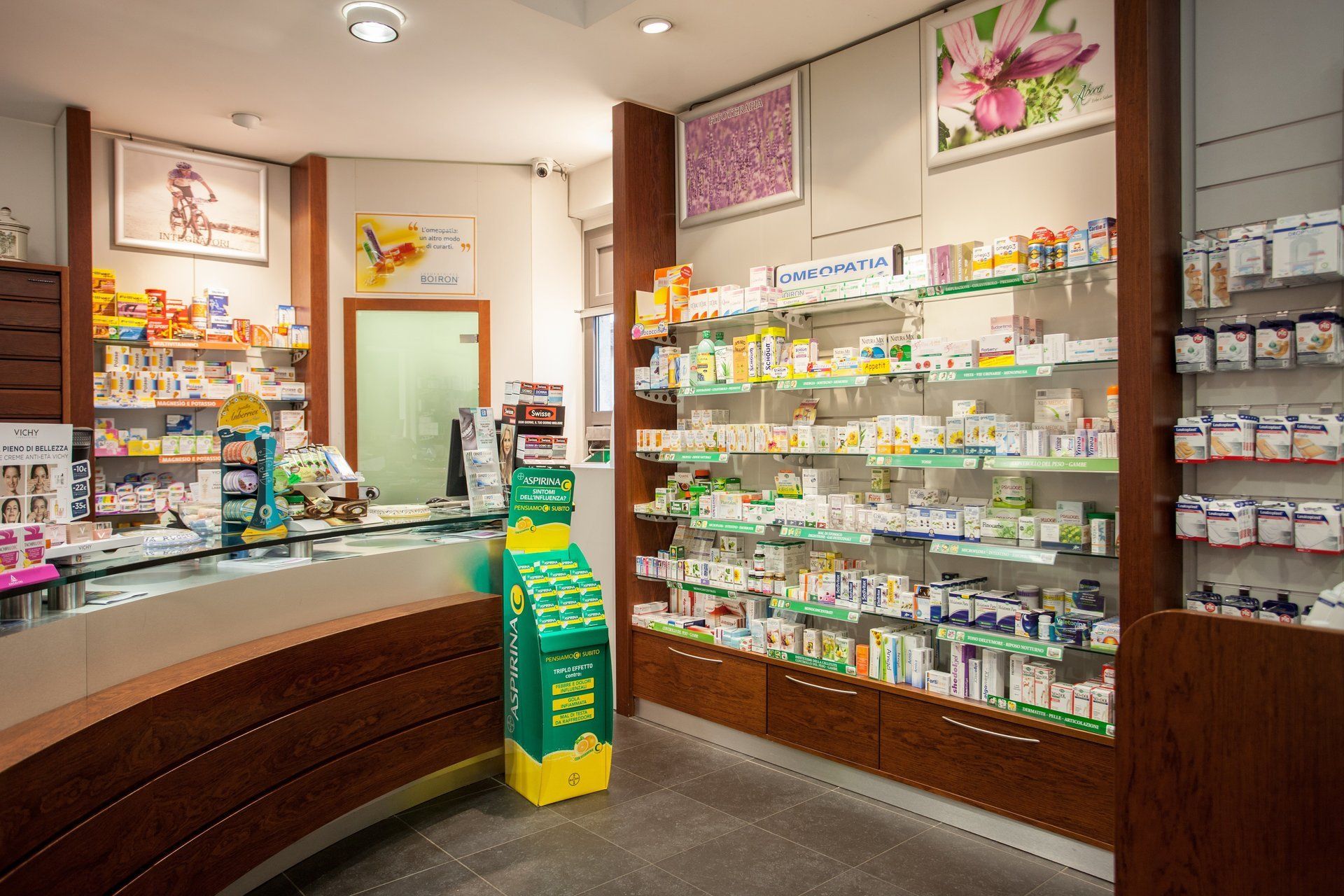 Il bancone di una farmacia con scaffali pieni di medicinali, un alto espositore verde per i prodotti e una luminosa illuminazione interna.