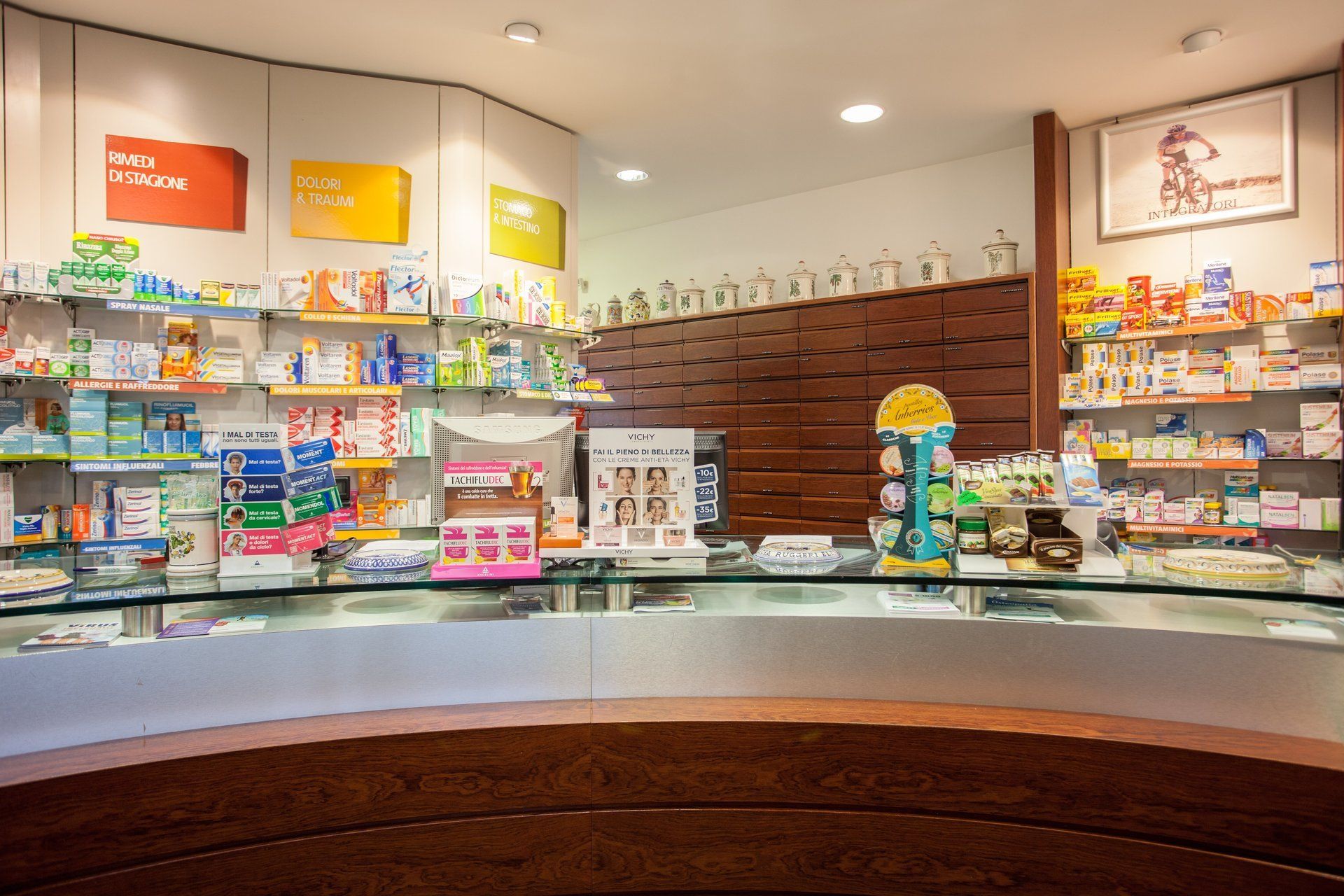 L'interno di una farmacia con i prodotti esposti sugli scaffali dietro un lungo bancone curvo in legno e vetro.