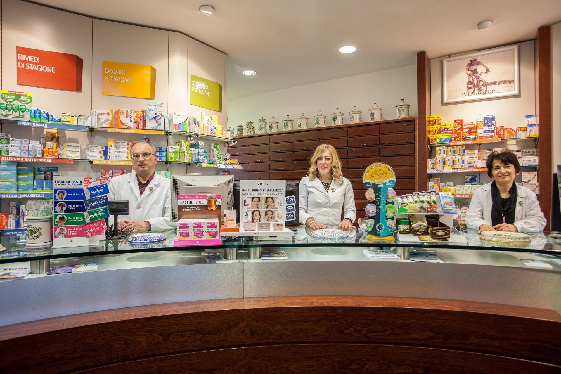 Tre farmacisti in camice bianco sono in piedi dietro il bancone di una farmacia, dove gli scaffali sono pieni di prodotti medicinali.