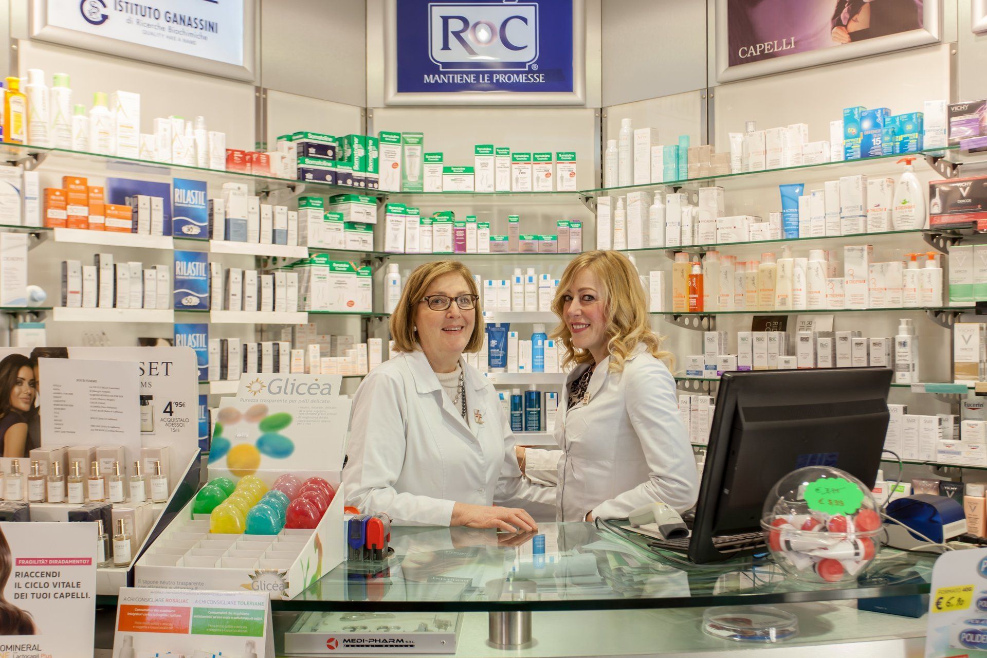 Due membri dello staff in camice bianco sono in piedi dietro il bancone di una farmacia, su cui sono disposti scaffali pieni di prodotti per la cura della pelle e per la salute.