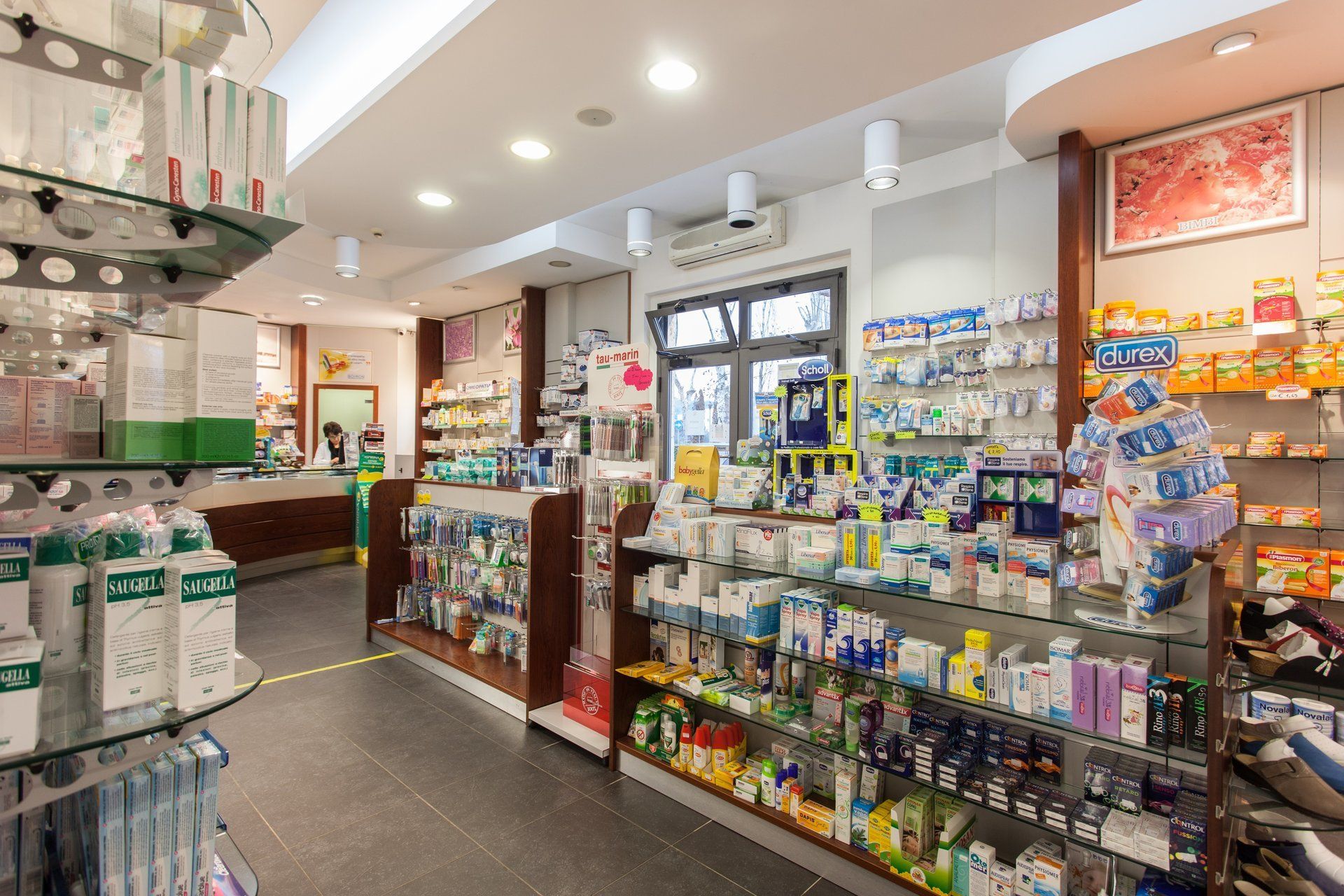 L'interno di una farmacia ben illuminata presenta corridoi pieni di vari prodotti per la salute e il benessere esposti su scaffali di vetro.