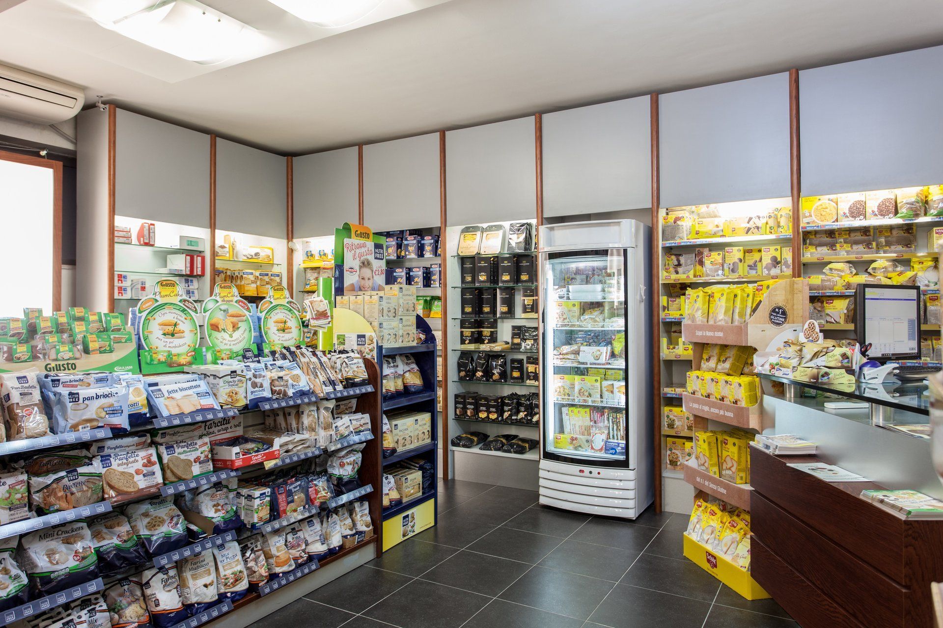 L'interno di un negozio ben illuminato presenta scaffali pieni di vari snack confezionati, un frigorifero bianco e una cassa.