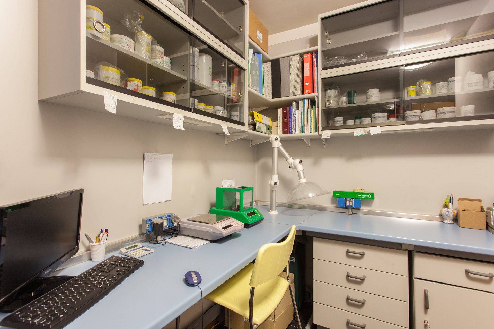 Un ufficio di laboratorio con un computer, bilance analitiche su una scrivania azzurra e scaffali pieni di forniture.