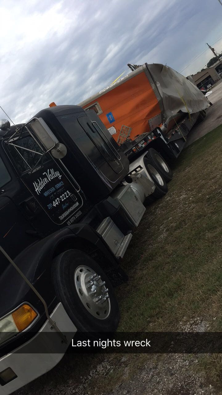 Black semi-truck on grass with orange tarp and wreckage, cloudy sky. 