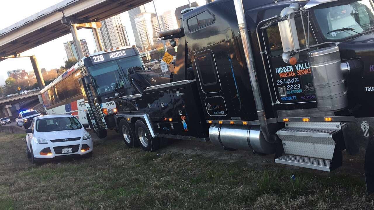 Tow truck removing a bus from a grassy area; white car, police car visible under overpass.