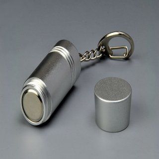 Contenedor de llavero cilíndrico plateado con su tapa quitada, apoyado sobre una superficie gris.