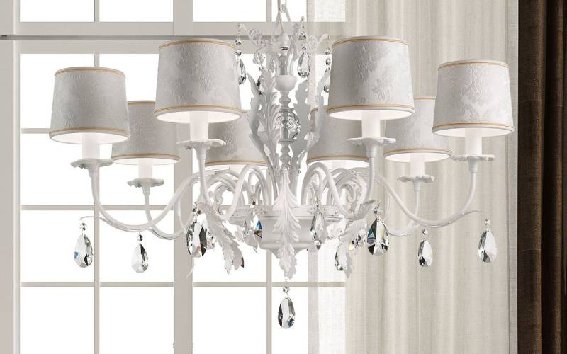 Lampadario bianco decorato con sei lampade, dettagli in cristallo e paralumi davanti a una finestra.