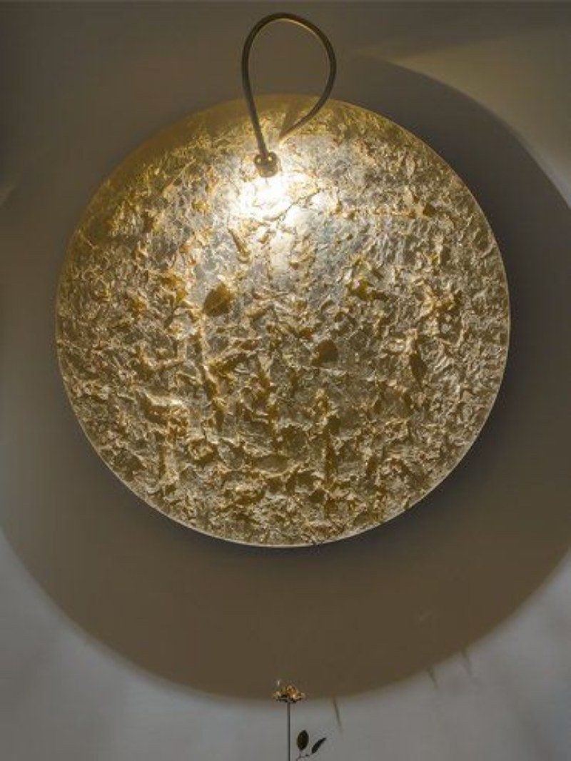 Lampada da parete con finitura dorata testurizzata, dotata di un anello per appenderla, che proietta un'ombra sulla parete sottostante.