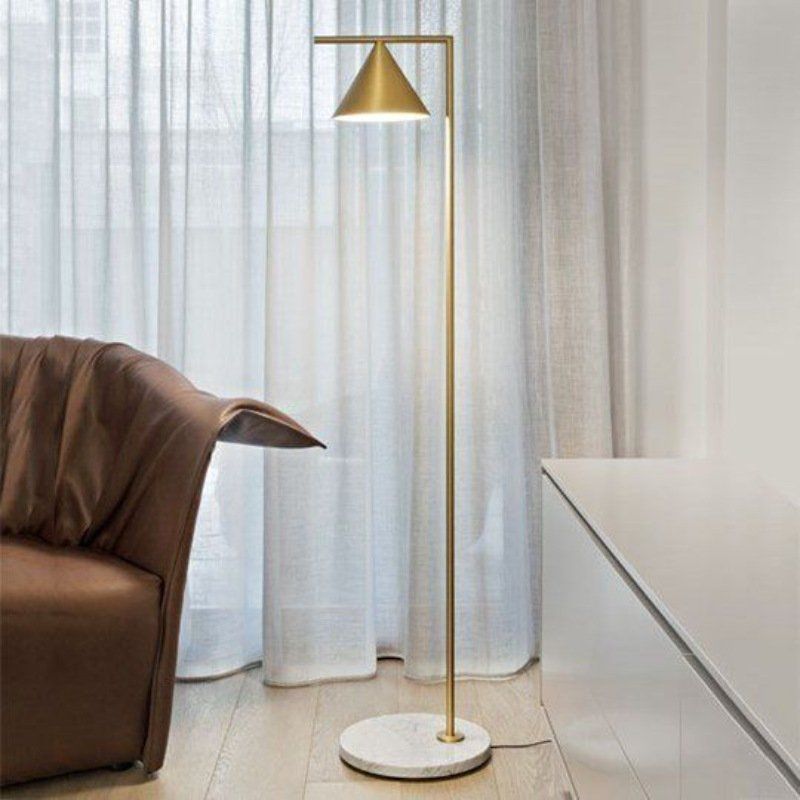 Lampada da terra dorata con paralume conico accanto a una poltrona in pelle e tende bianche.