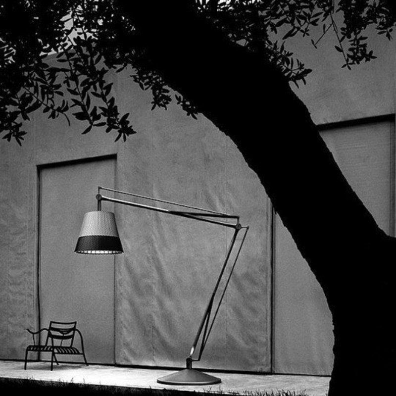 Scena esterna in bianco e nero: una lampada alta e articolata davanti a un muro, con un tronco d'albero in primo piano e una sedia a sinistra.