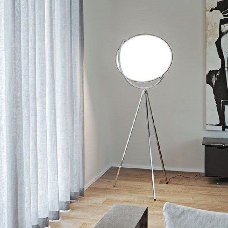 Lampada da terra a treppiede argentata con una grande e luminosa luce circolare bianca, vicino a tende bianche.