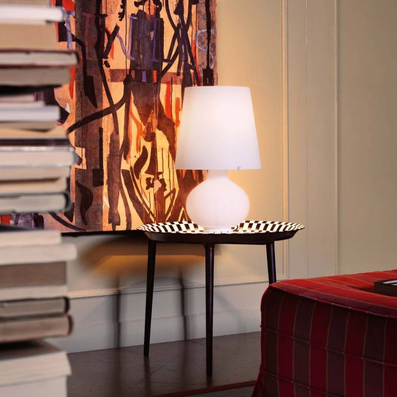 Lampada bianca su un tavolino con piano decorato, accanto a una pila di libri e un divano rosso.