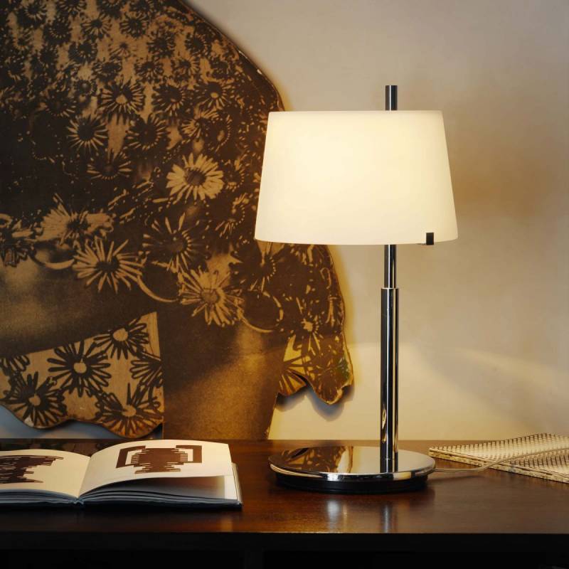 Lampada da scrivania con paralume bianco su un tavolo, con un libro aperto e un'opera d'arte floreale sullo sfondo.