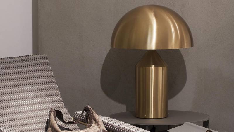 Lampada a cupola dorata su un tavolino, accanto a una sedia decorata e a una parete grigia.