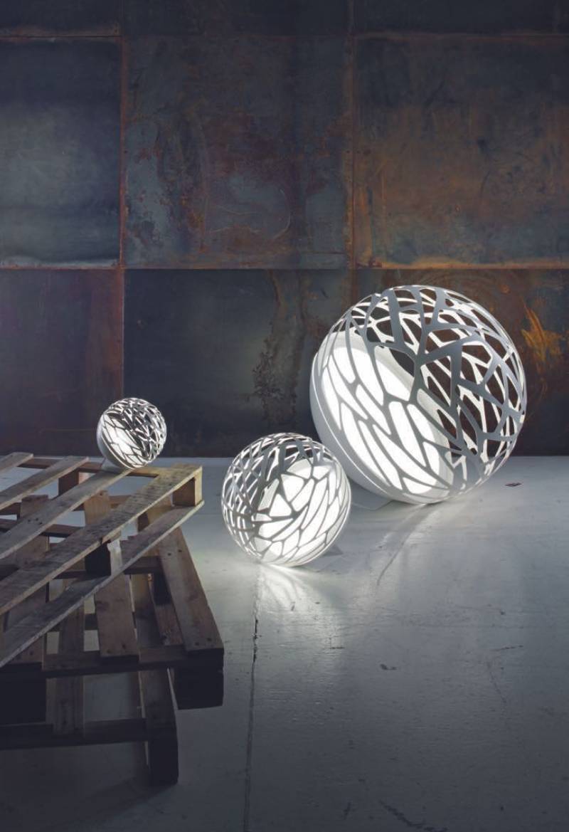 Quattro sculture sferiche illuminate con intricati ritagli, appoggiate contro una parete di metallo arrugginito e un pallet di legno.