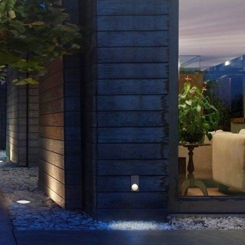 Esterno di un edificio moderno con illuminazione soffusa. Il rivestimento in legno e una finestra rivelano un interno ricco di piante.