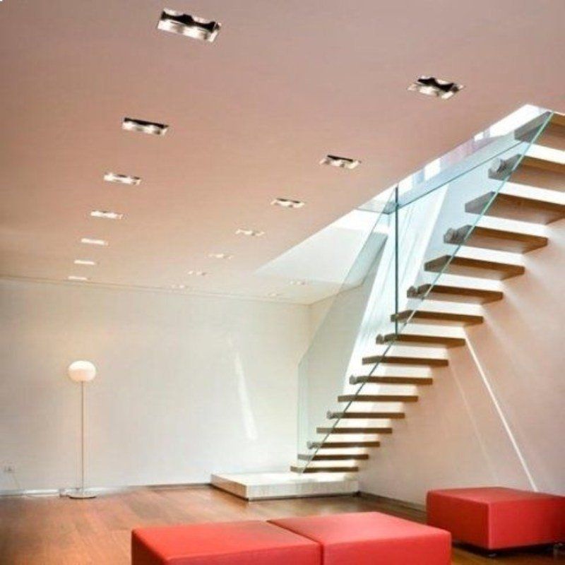 Interni moderni con scale sospese, ringhiera in vetro e faretti a incasso nel soffitto. Pouf rossi e una lampada da terra.