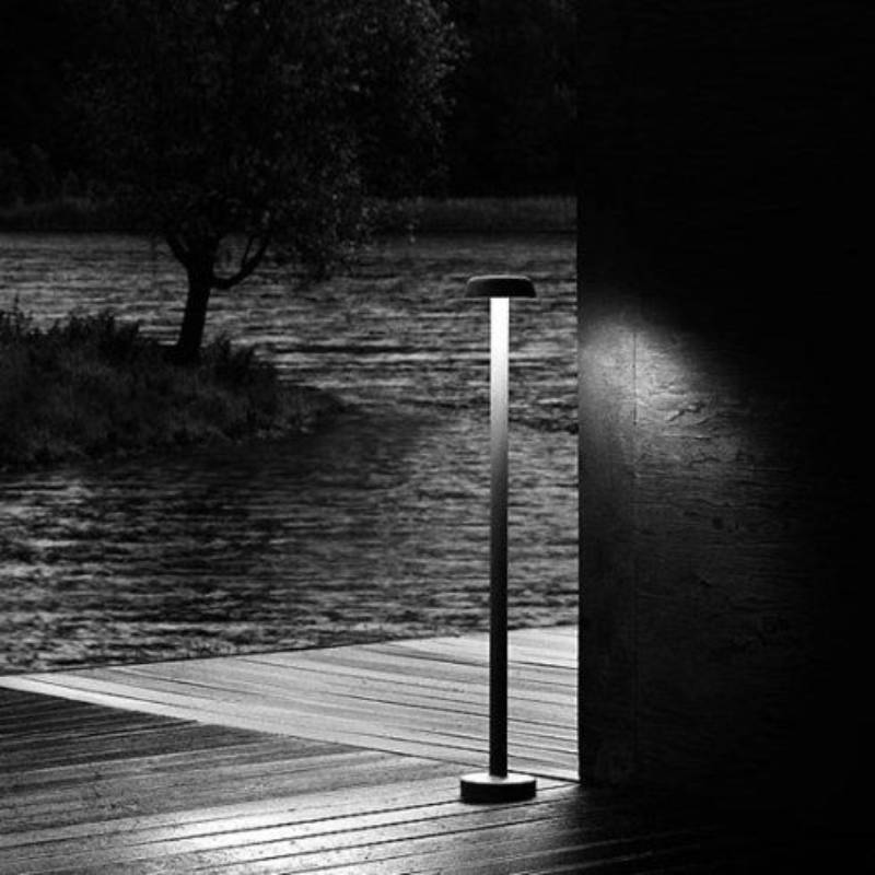 Foto in bianco e nero di una lampada da esterno accesa su un ponte di legno, vicino all'acqua e con un albero sullo sfondo.