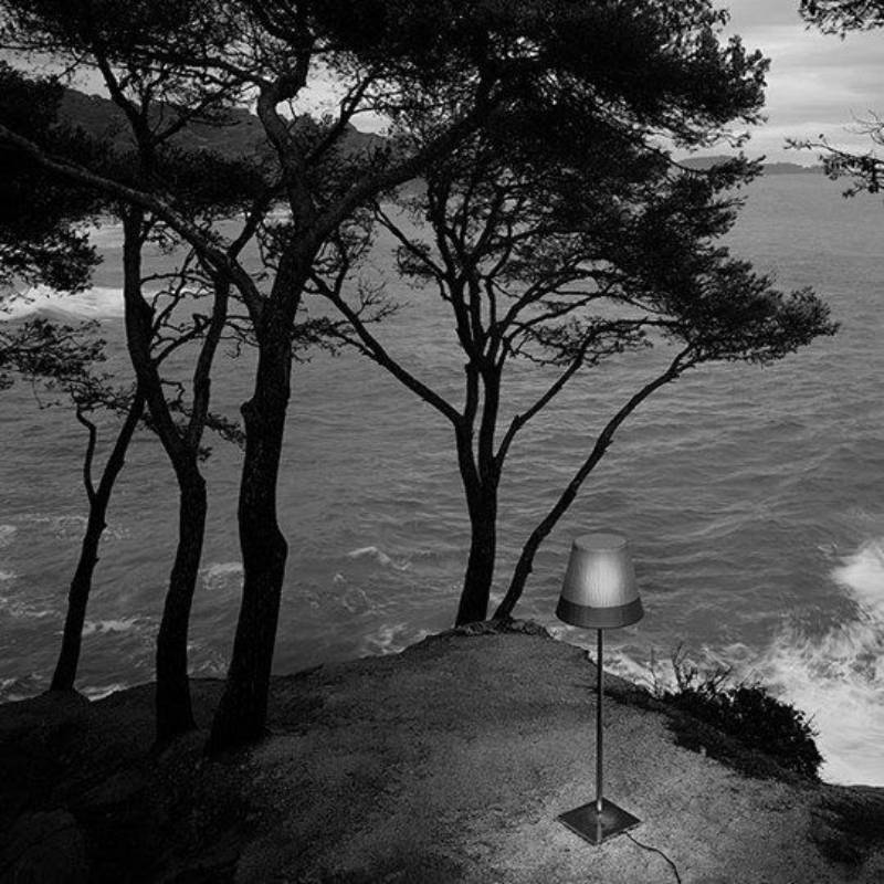 Foto in bianco e nero: una lampada è posta su una scogliera che domina un oceano ondoso; sullo sfondo si vedono degli alberi.