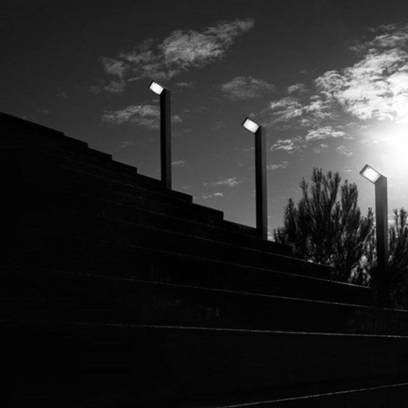 Immagine in bianco e nero dei gradini di uno stadio con tre luci brillanti sullo sfondo di un cielo parzialmente nuvoloso.
