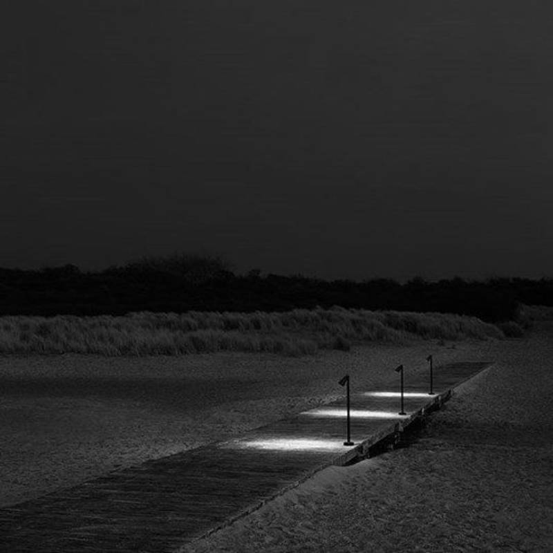 Una passerella notturna con luci accese che conduce alle dune e a un orizzonte scuro.