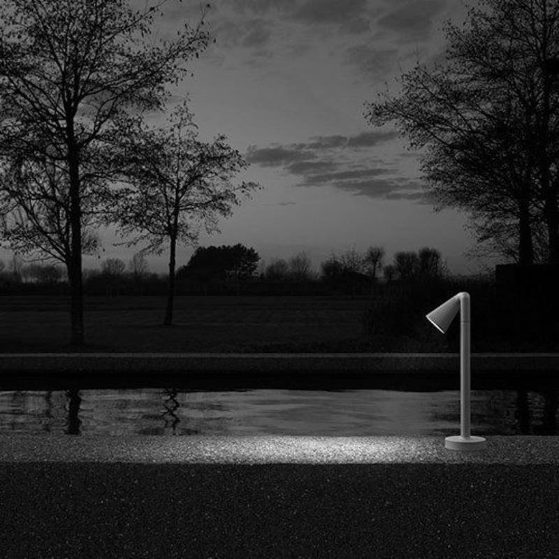 Scena notturna: una moderna luce angolata illumina un marciapiede in cemento accanto a uno specchio d'acqua. Alberi sullo sfondo.