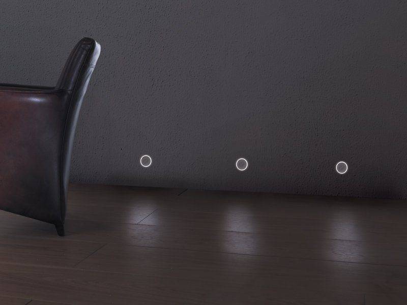 Una stanza buia con una sedia, luci da terra che illuminano un pavimento in legno, contro una parete scura.