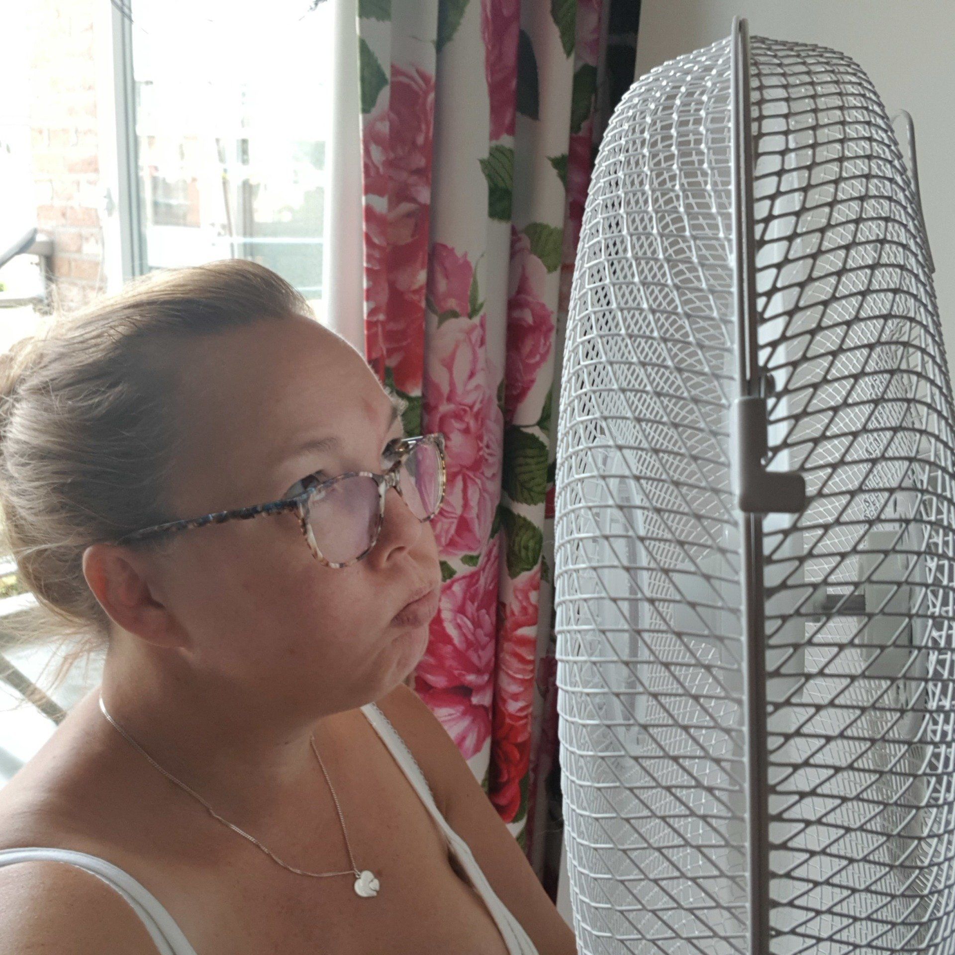 Een vrouw met een bril kijkt naar een staande ventilator. Ze heeft een lichte huid en een lichtroze bloemengordijn.
