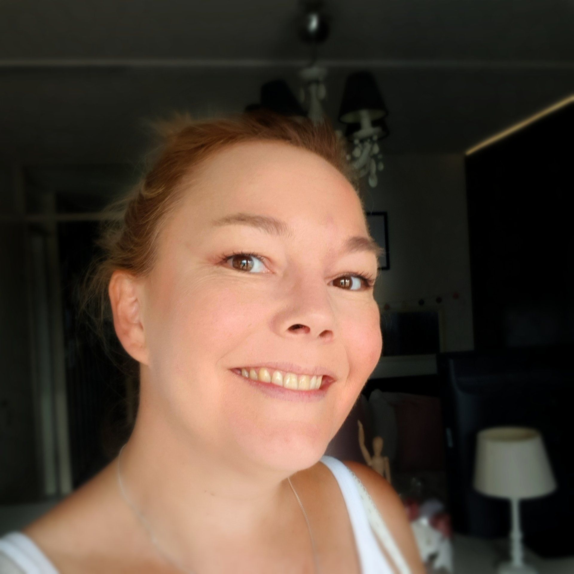 Een vrouw met een lichte huid glimlacht naar de camera. Ze heeft lichtrood haar en draagt ​​een wit tanktopje. Donker vanbinnen.