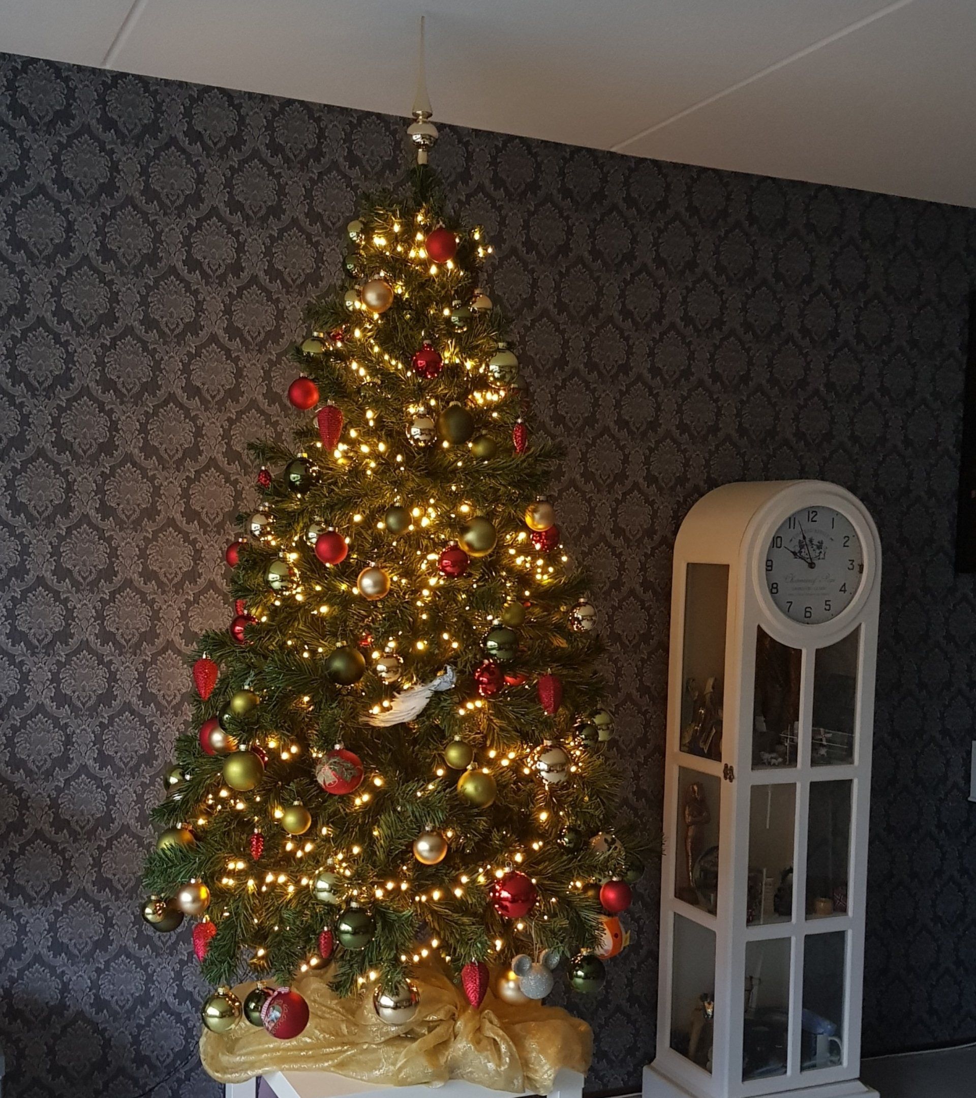 Kerstboom versierd met lichtjes en ornamenten voor een muur met patroon, naast een witte staande klok.