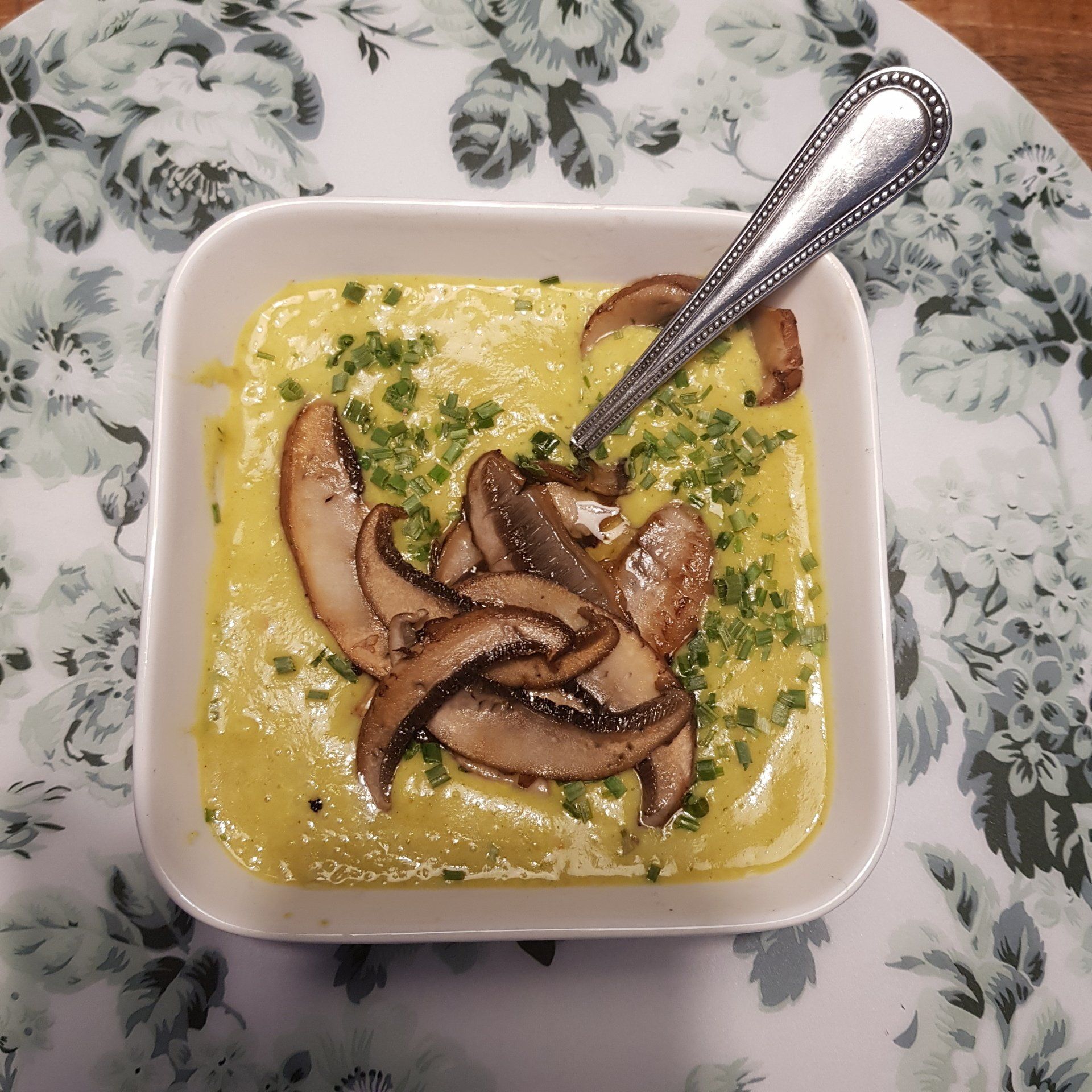 Romige gele soep in een witte vierkante kom, gegarneerd met plakjes champignons en bieslook en een zilveren lepel.